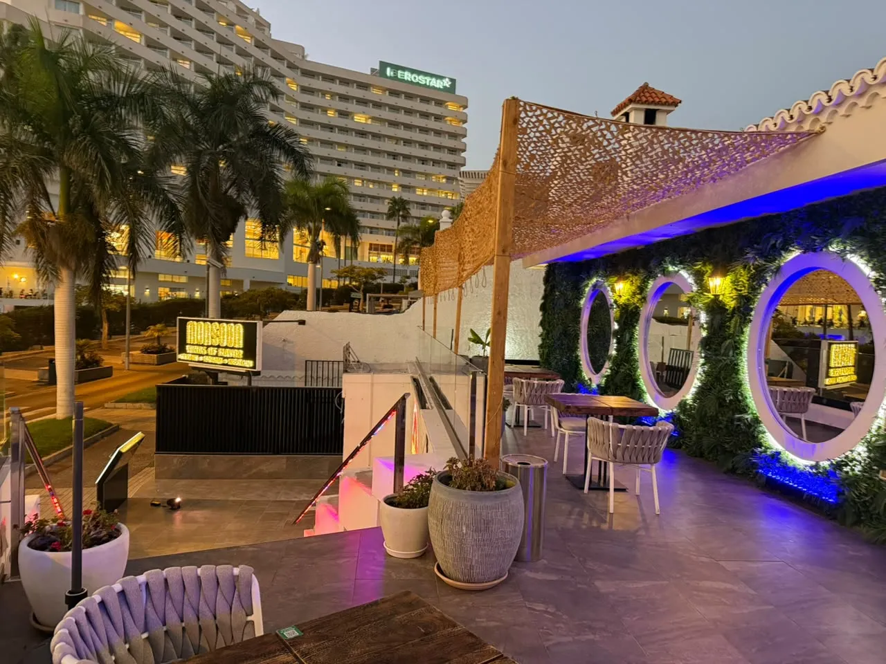 Terrace seating beside circular illuminated mirrors and greenery at Monsoon Tenerife